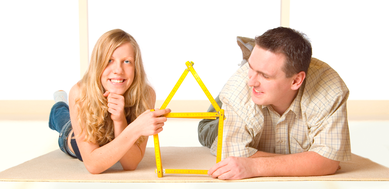 Pärchen auf Fußboden liegend in unmöblierter Wohnung hält ein Haus geformt aus einem Zollstock