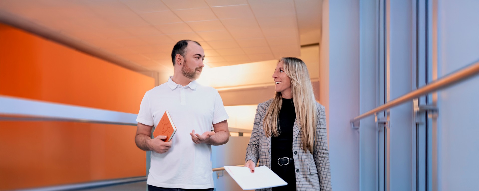 Kollege geht mit Kollegin den Flur entlang und bespricht etwas.