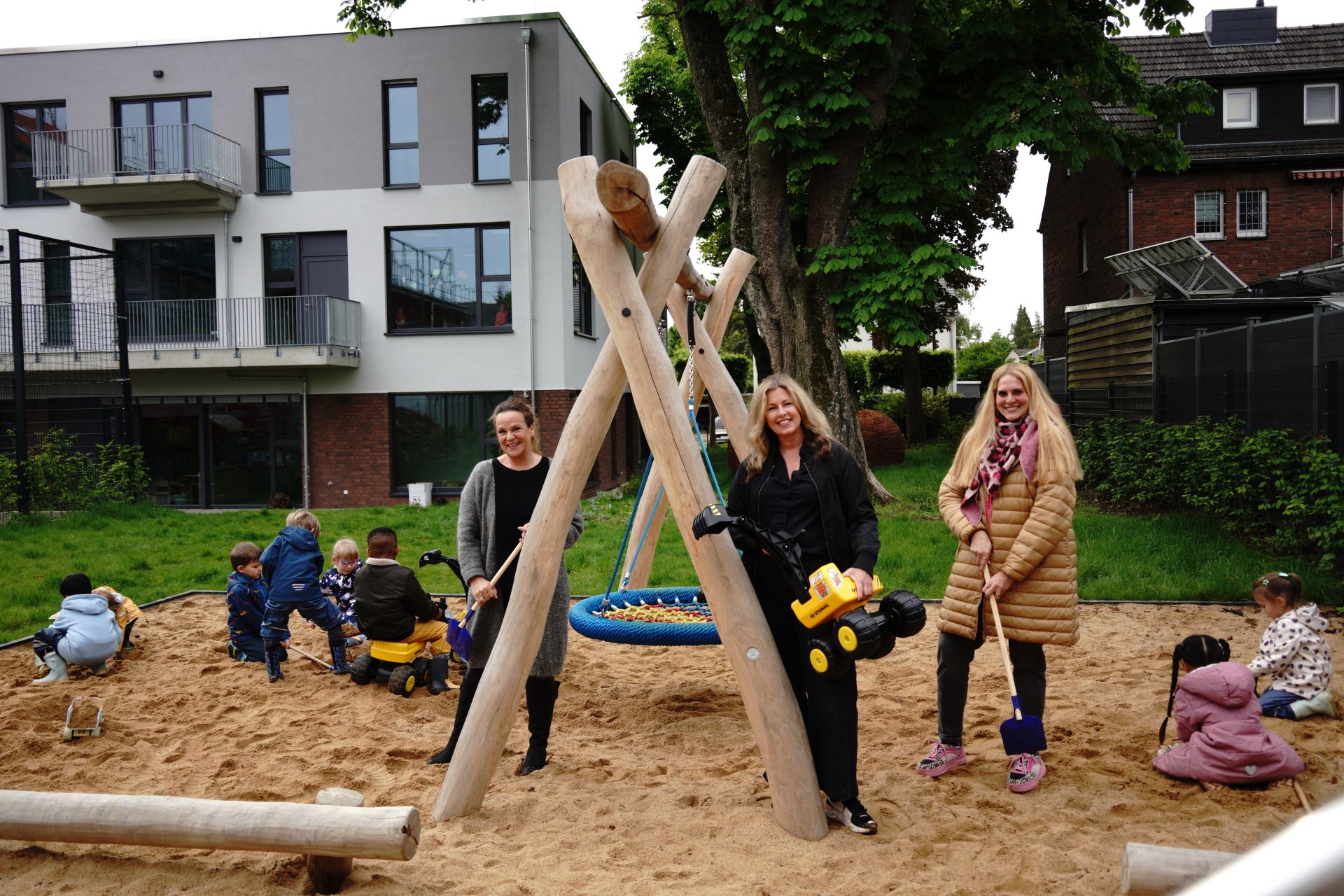 Sandkasten mit Nestschaukel, in dem Kinder mit Sandspielzeug spielen und nita Vondermaßen, Fachbereich KiTa und Kindertagespflege, Michaela Sprenger, STAWAG und Nadia von Thenen, stellvertretende KiTa-Leiterin dabei stehen