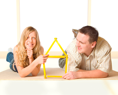 Pärchen auf Fußboden liegend in unmöblierter Wohnung hält ein Haus geformt aus einem Zollstock